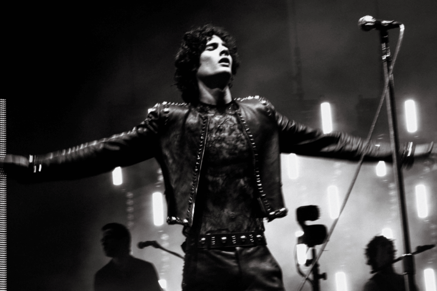 Close-up black and white shot of Sombr performing live on stage at Coachella 2026. He is wearing a studded black leather jacket, with arms outstretched and a passionate expression, backlit by dramatic stage lighting.