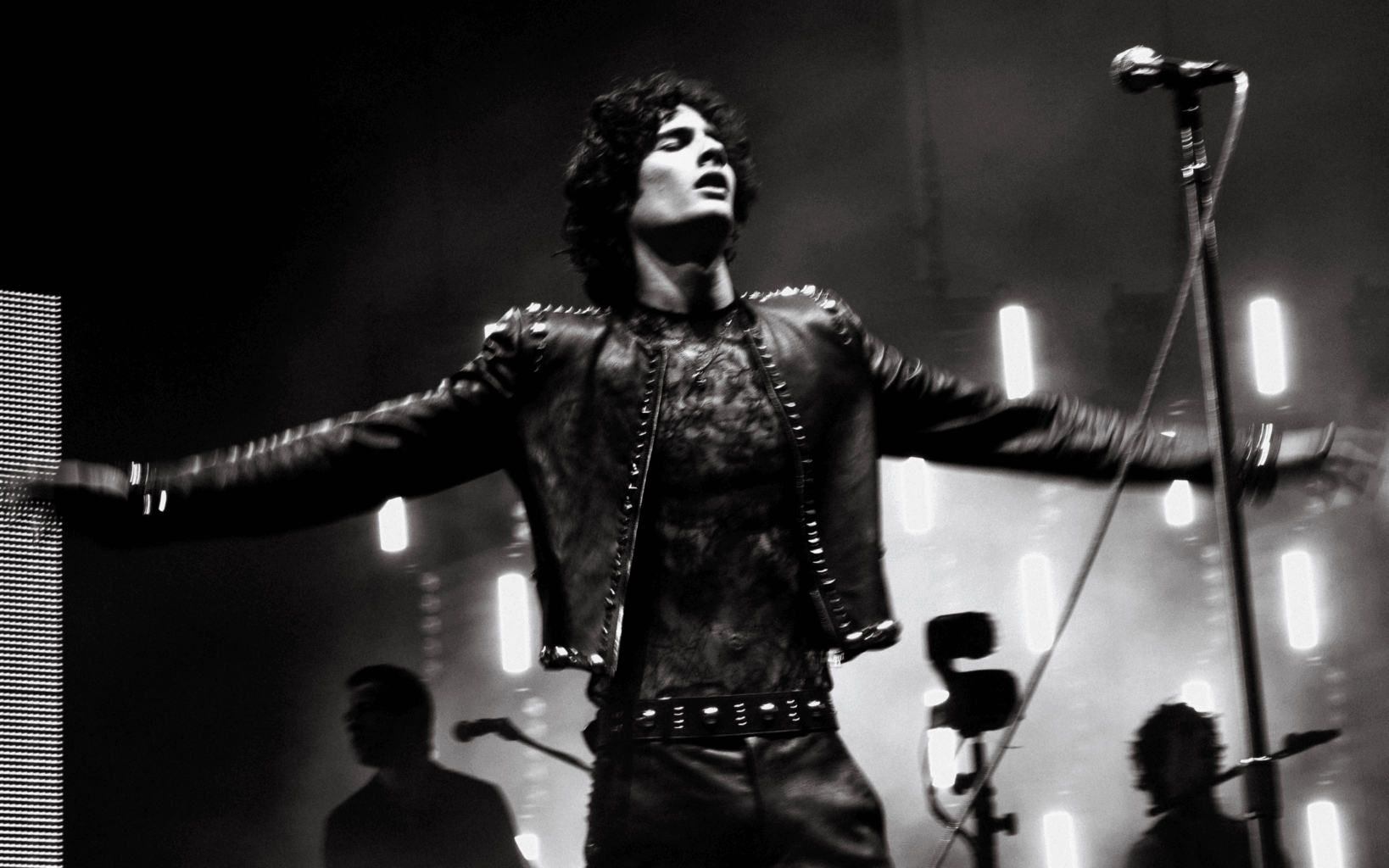 Close-up black and white shot of Sombr performing live on stage at Coachella 2026. He is wearing a studded black leather jacket, with arms outstretched and a passionate expression, backlit by dramatic stage lighting.